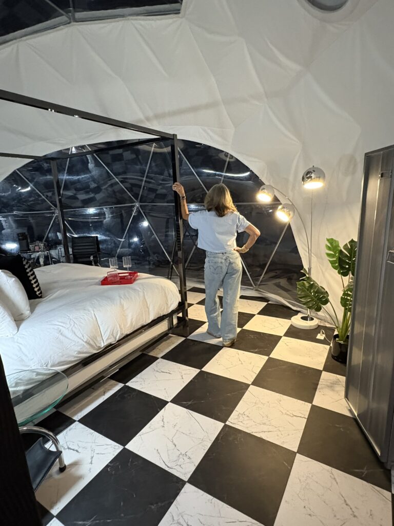 Interior of Starman lodging at All Is Well St. Jo, featuring a woman standing near a bed, modern decor, checkered floor, and large windows, highlighting unique accommodation experience.