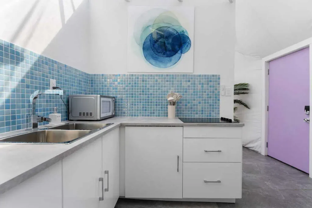Modern kitchen interior in Escape Velocity Dome featuring blue tiled backsplash, stainless steel sink, microwave, and colorful wall art, emphasizing unique lodging experience.
