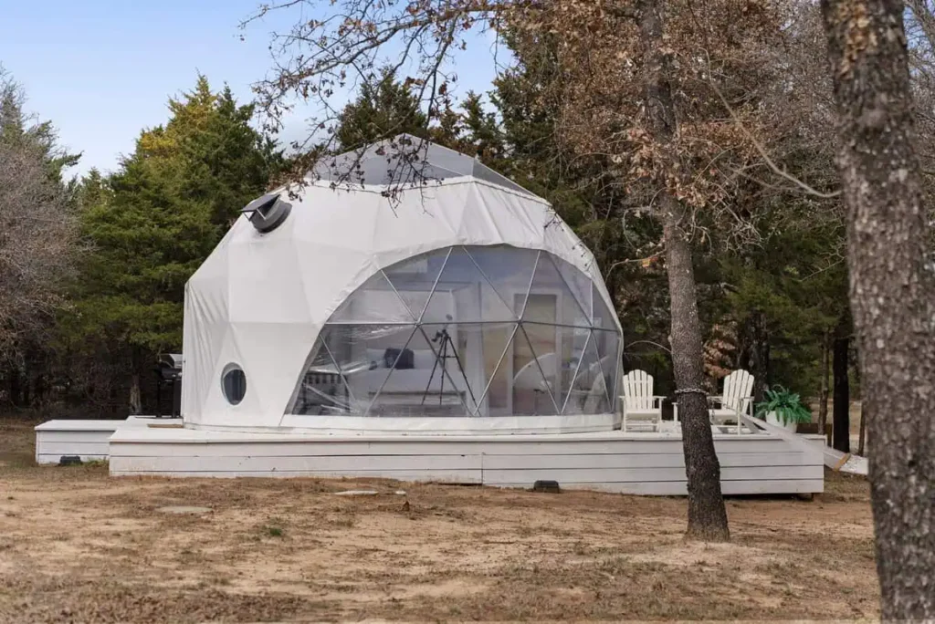 Unique geodesic dome accommodation at Escape Velocity, surrounded by trees, featuring a spacious deck with white chairs, ideal for a scenic getaway.