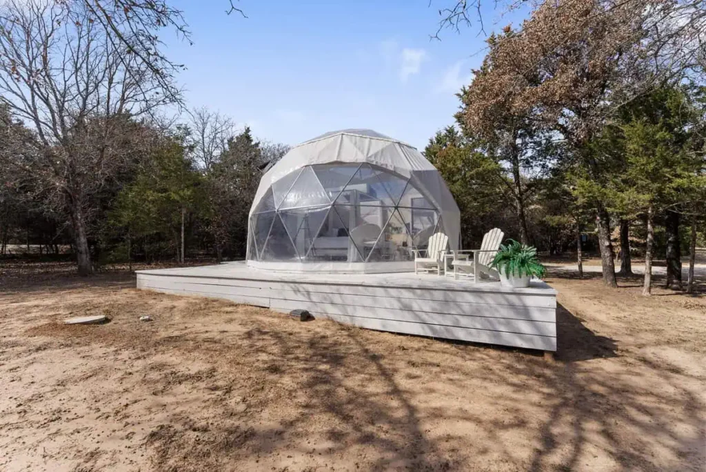 Escape Velocity dome accommodation with geodesic design, wooden deck, and seating area, surrounded by trees and nature, offering a unique lodging experience.