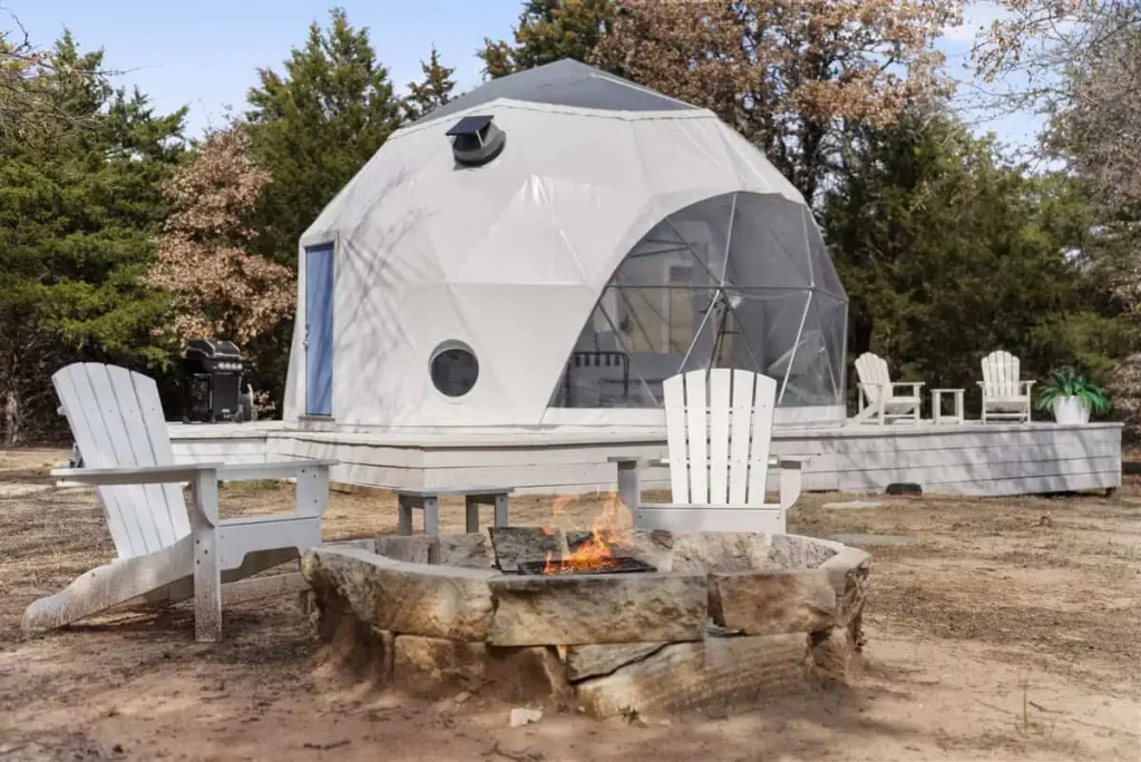 Geodesic dome accommodation with fire pit and Adirondack chairs, surrounded by natural scenery, showcasing the unique Escape Velocity lodging experience.