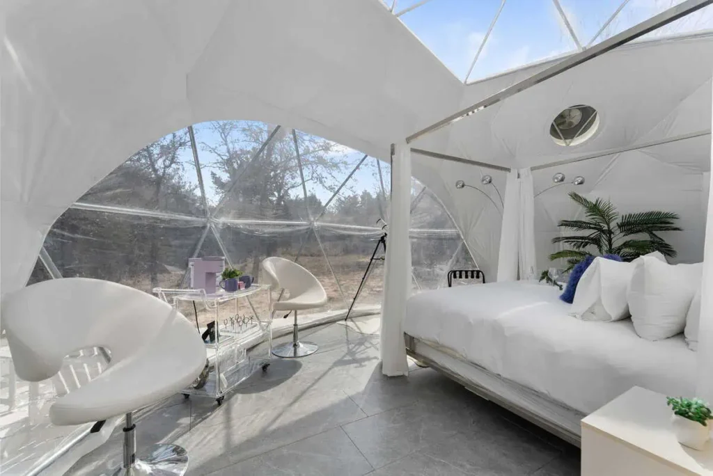 Interior of Escape Velocity dome with a cozy bed, modern seating, and large windows showcasing nature, emphasizing unique geodesic lodging experience.