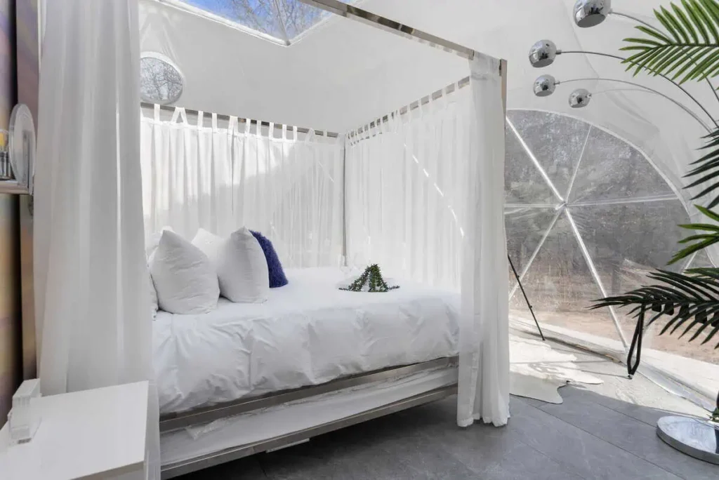 Interior of Escape Velocity Dome featuring a cozy bed with white drapes, decorative pillows, and natural light illuminating the space, emphasizing a unique and comfortable lodging experience.