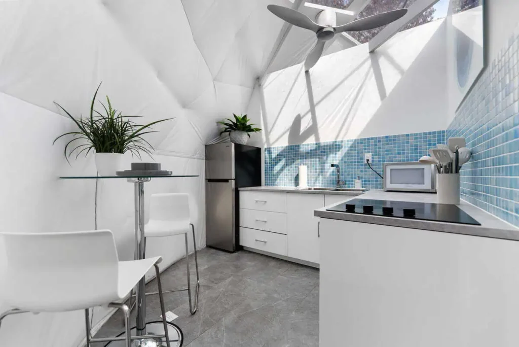 Modern kitchen interior of the Escape Velocity dome, featuring a stainless steel refrigerator, microwave, and blue tiled backsplash, complemented by a glass dining table and white chairs.