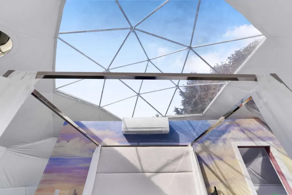 Interior view of the Escape Velocity dome featuring a geodesic ceiling with a clear sky, air conditioning unit, and colorful wall art, highlighting unique lodging experience at All is Well St. Joseph.