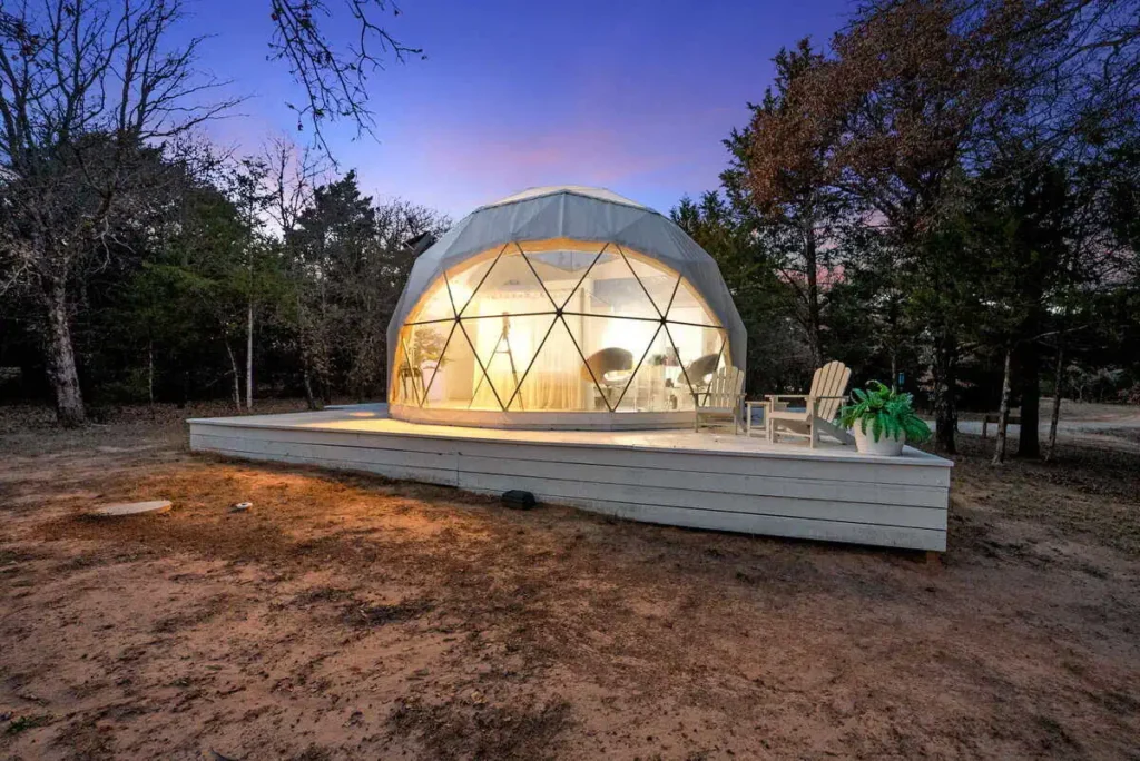 Escape Velocity dome accommodation at twilight, featuring a geodesic design, illuminated interior, and outdoor seating area, surrounded by trees in a scenic setting.