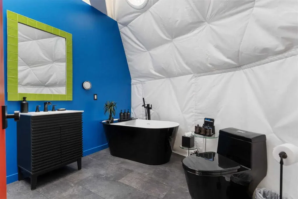 Modern bathroom interior in a dome accommodation featuring a blue wall, black fixtures, a freestanding bathtub, and a green-framed mirror, highlighting unique lodging at All Is Well Resort.