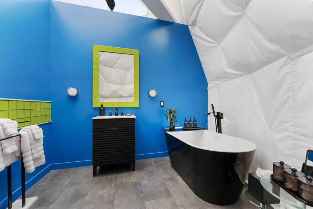 Colorful bathroom interior in dome accommodation, featuring a blue wall, green-accented mirror, modern black freestanding bathtub, and stylish amenities for guests at All Is Well Resort.