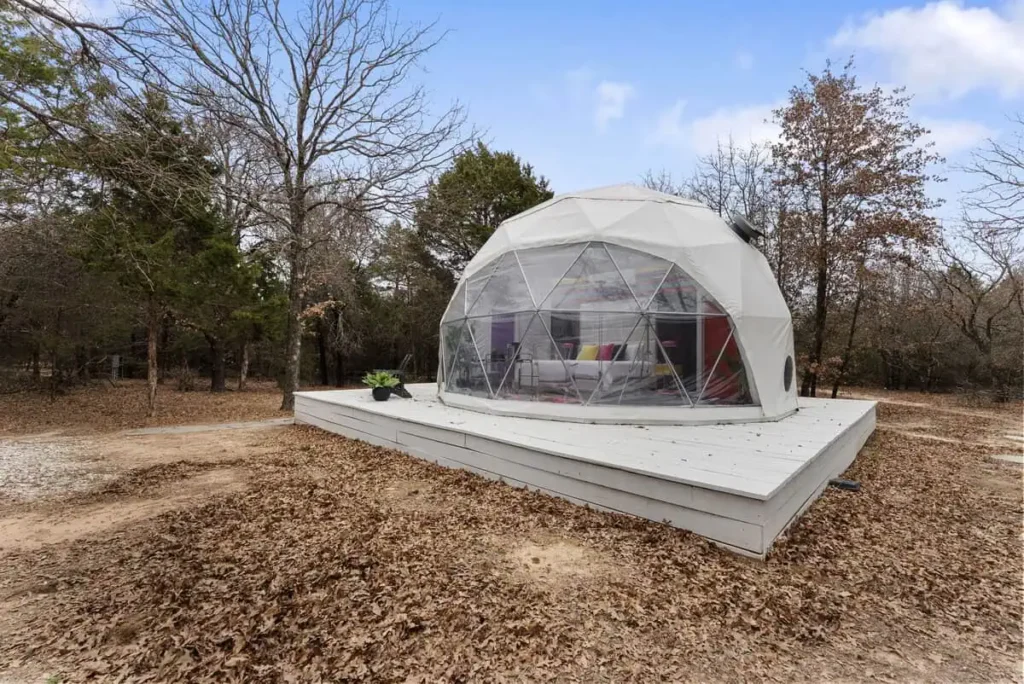 Dome-shaped accommodation at All Is Well Resort, featuring a transparent exterior, surrounded by trees and fallen leaves, emphasizing unique lodging experience in nature.