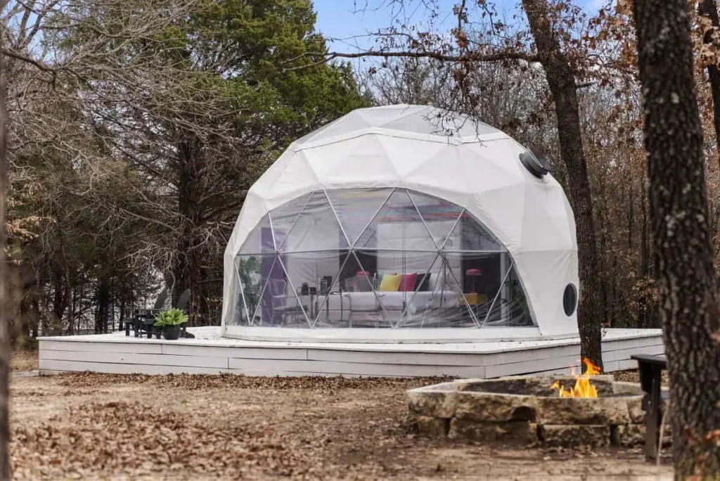 Dome accommodation at All Is Well Resort featuring large windows, colorful interior, and outdoor fire pit surrounded by trees.