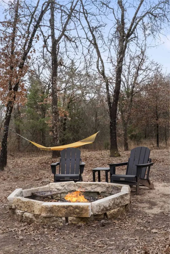 Outdoor fire pit with stone seating, two black Adirondack chairs, and a yellow hammock in a natural setting at All Is Well Resort.