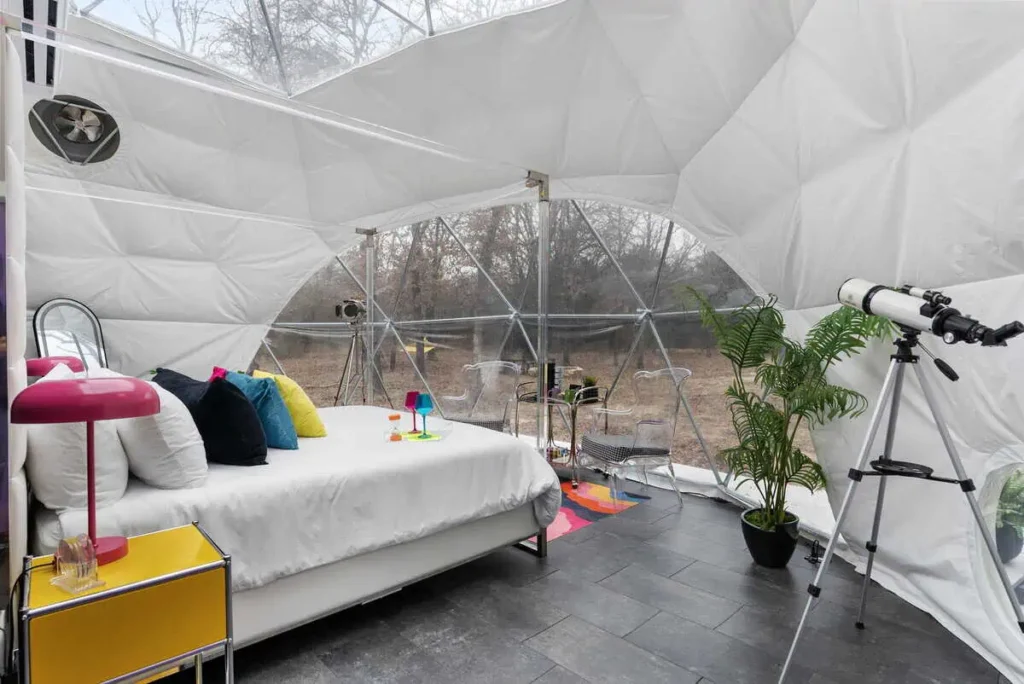 Interior of a modern dome accommodation featuring a bed with colorful pillows, a vibrant lamp, a telescope, and a small table with drinks, set in a natural landscape.