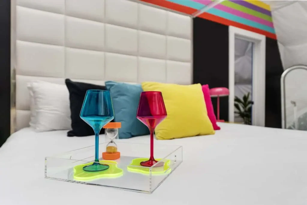 Colorful wine glasses on a tray with a sand timer, set against a vibrant, modern interior featuring decorative pillows and a stylish backdrop, highlighting the unique ambiance of the Mod Squad accommodation at All Is Well Resort.