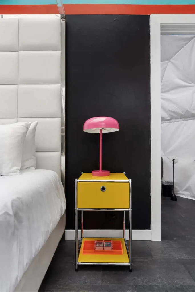 Colorful bedside table with yellow drawer and pink lamp beside a modern bed in the 'Mod Squad' accommodation at All Is Well Resort.