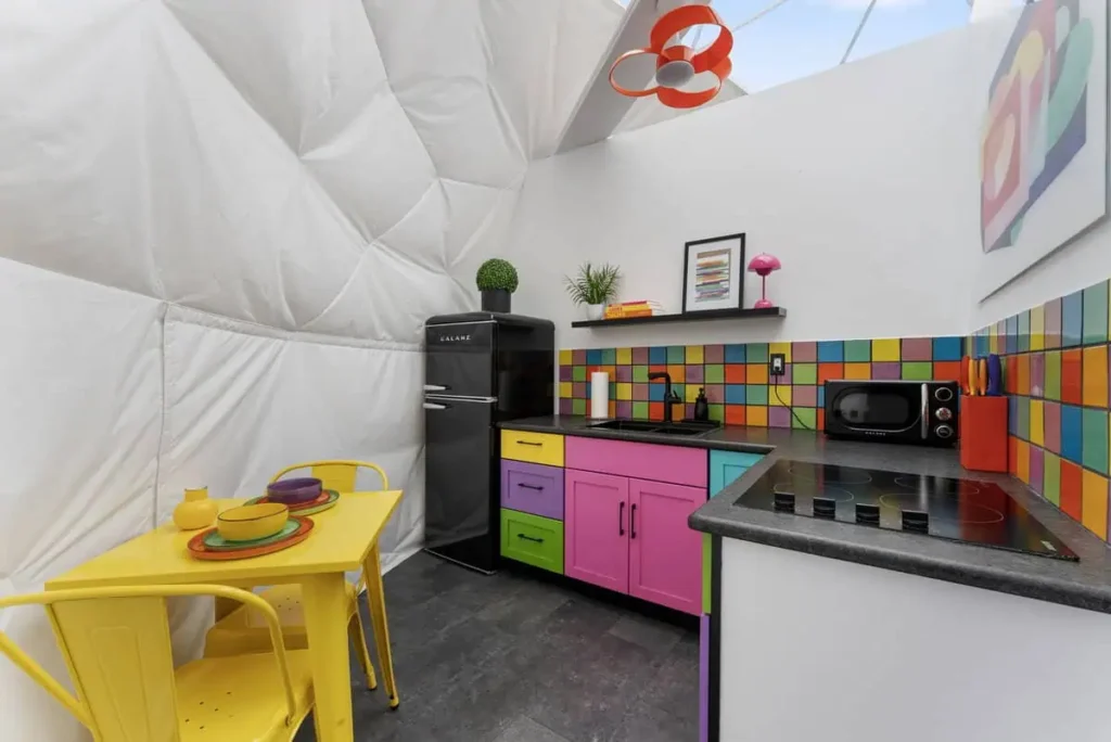 Colorful kitchen in 'Mod Squad' dome accommodation at All Is Well Resort, featuring a black refrigerator, vibrant cabinetry, and dining table with plates.