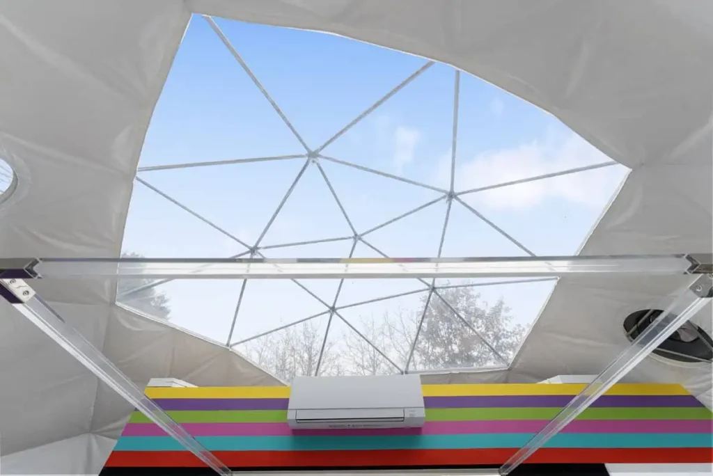 Interior view of a colorful dome accommodation at All Is Well Resort, featuring a geometric skylight, air conditioning unit, and vibrant striped counter, showcasing unique lodging experience in nature.