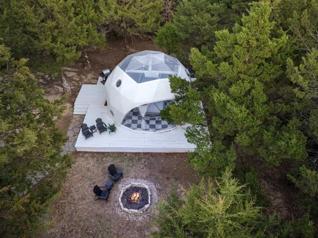Aerial view of the Starman dome accommodation at All Is Well St. Jo, featuring a fire pit, wooden deck, and surrounding trees, highlighting unique lodging amenities for a relaxing stay.