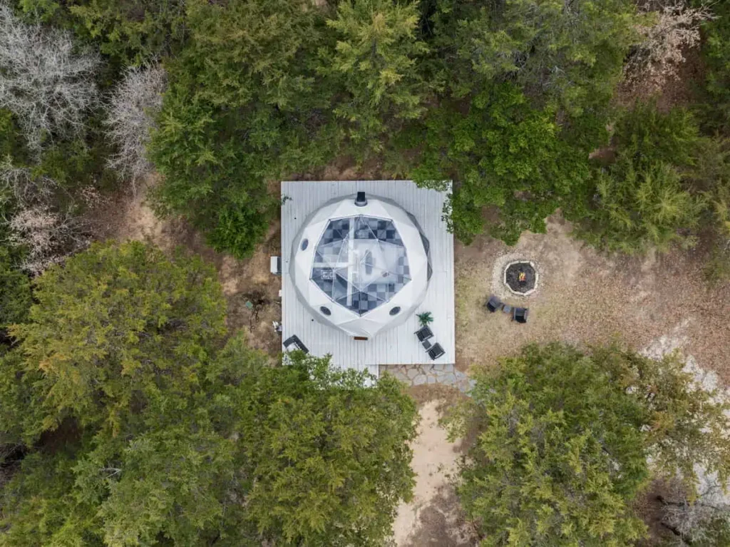Aerial view of Starman dome lodging surrounded by trees, featuring a wooden deck, outdoor seating, and a fire pit in a serene natural setting.