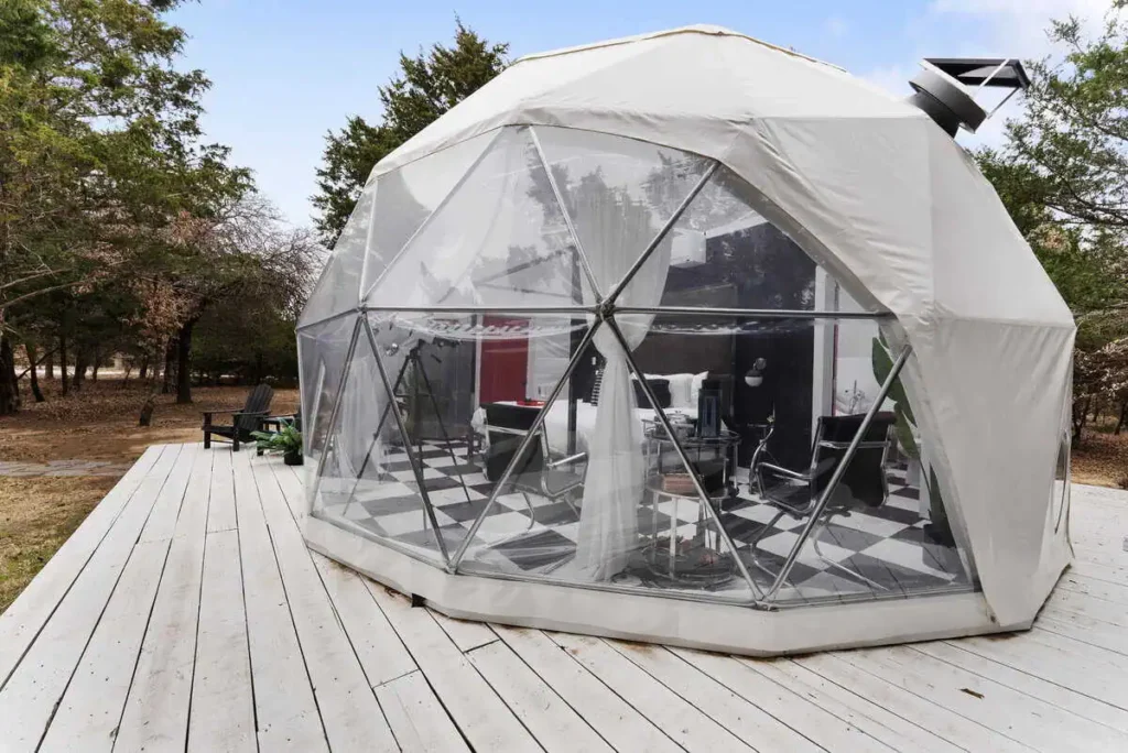 Dome-shaped accommodation at Starman lodging, featuring transparent walls, modern interior design, and a wooden deck surrounded by trees, highlighting unique lodging experience at All Is Well St. Jo.