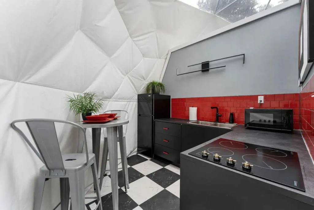 Modern kitchen interior in Starman lodging, featuring black cabinetry, red tile backsplash, countertop stove, microwave, refrigerator, and dining table with red plates, surrounded by a unique dome structure.