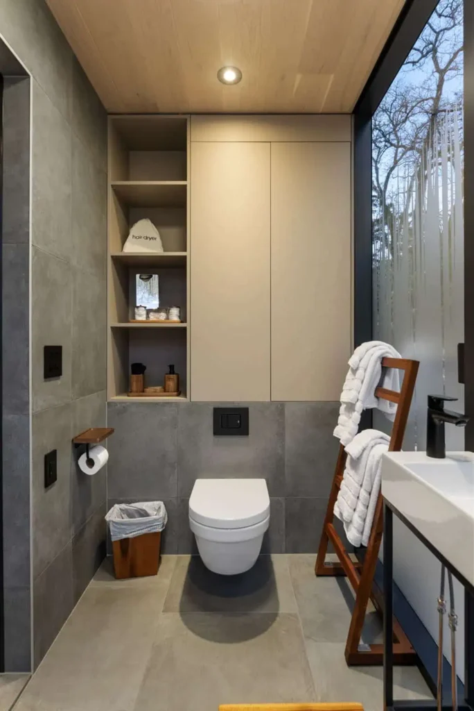 Modern bathroom design featuring a wall-mounted toilet, sleek sink, and wooden shelving with toiletries, towels, and a hairdryer, emphasizing the stylish amenities of the Cosmic Cube accommodation at All Is Well.