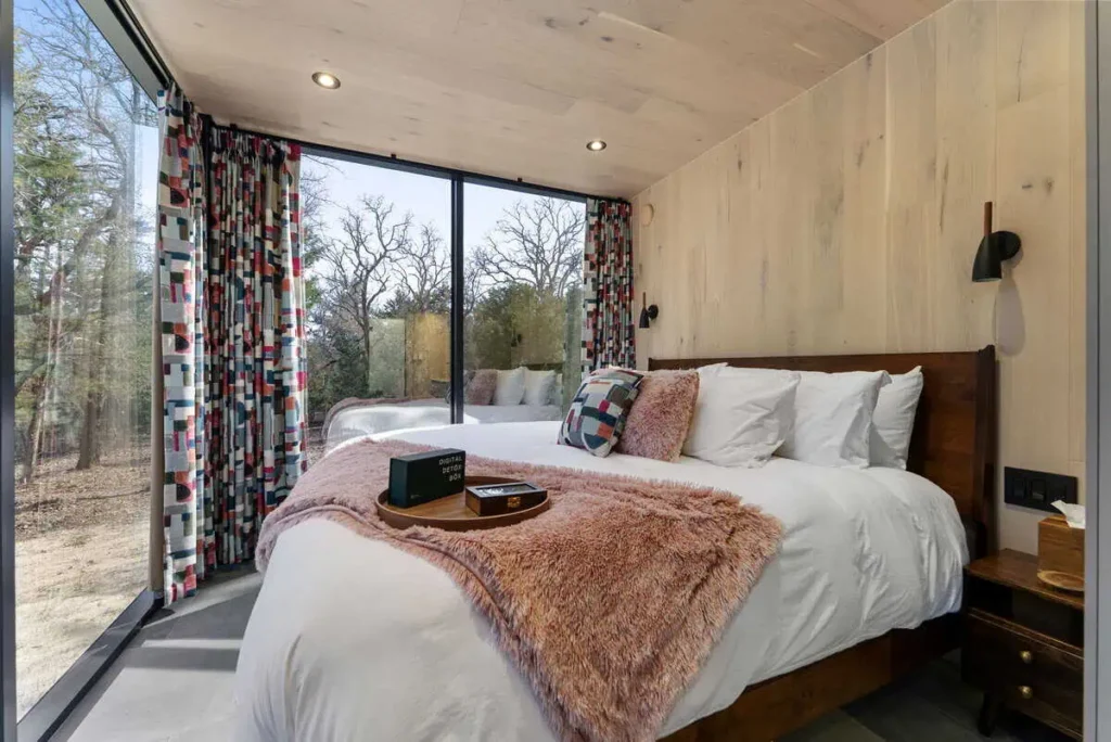 Cozy bedroom in the Cosmic Cube accommodation featuring a large bed, colorful patterned curtains, and large windows overlooking nature, emphasizing a unique lodging experience at All Is Well.