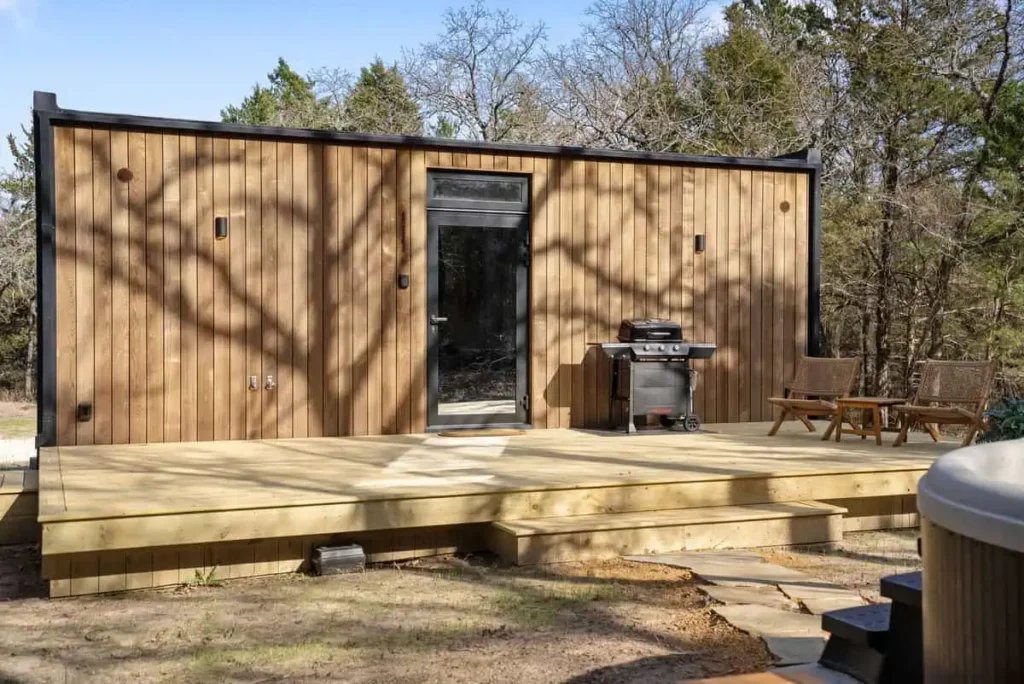 Cosmic Cube lodging exterior featuring wooden walls, deck, outdoor grill, and seating area, surrounded by trees.