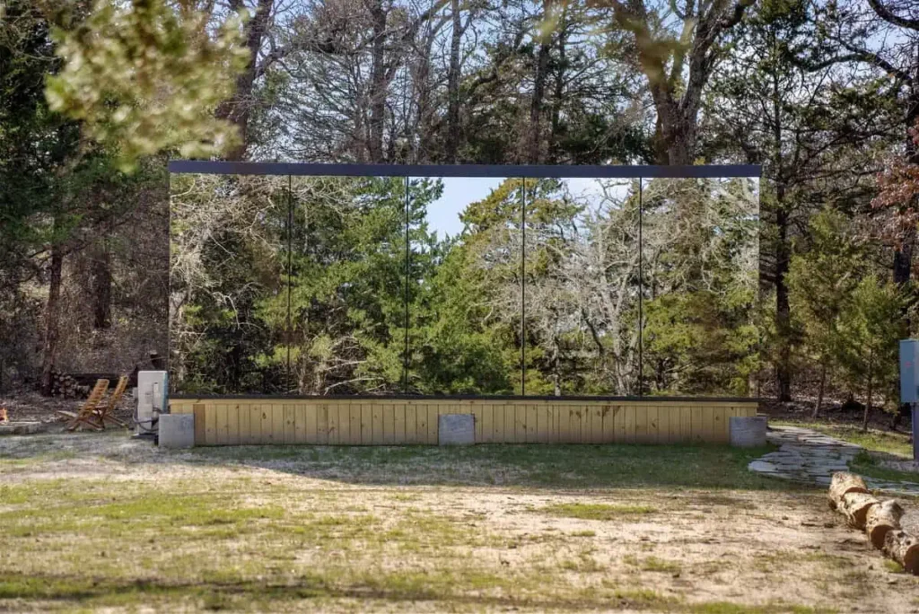 Mirror House exterior featuring a large reflective glass surface surrounded by trees, showcasing a unique architectural design for the Cosmic Cube lodging experience at All Is Well.