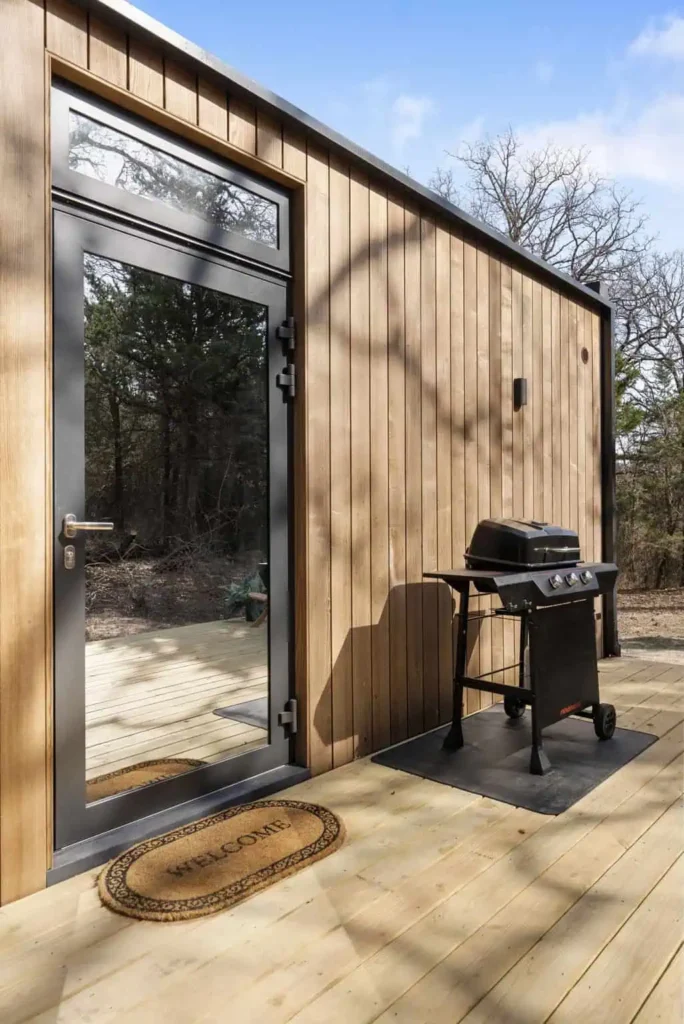 Exterior view of the Cosmic Cube accommodation featuring a welcoming entrance, wooden paneling, and a propane grill on the deck, highlighting unique lodging experience by All Is Well.