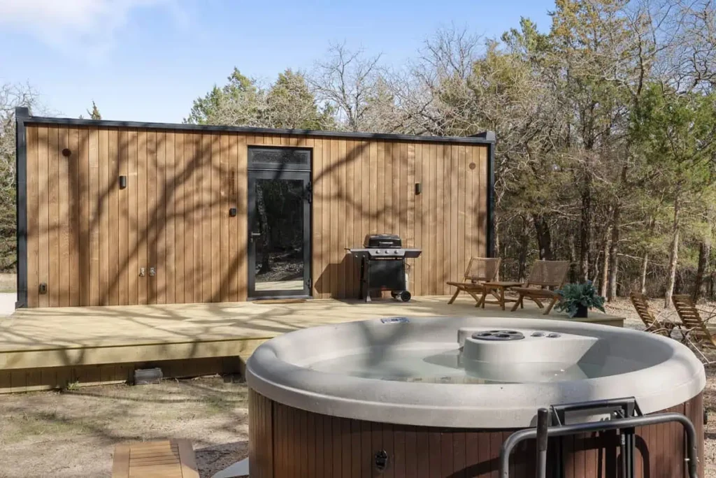 Hot tub in front of modern wooden cabin with outdoor seating area, surrounded by trees, showcasing amenities for a relaxing stay at Cosmic Cube accommodation by All Is Well.