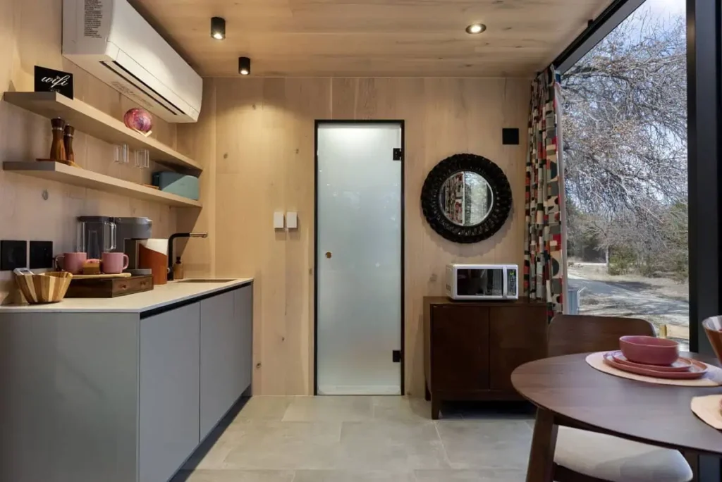 Modern kitchen interior of the Cosmic Cube accommodation featuring a microwave, mini fridge, Keurig coffee machine, and dining area with colorful tableware, showcasing a stylish and functional design for guests at All Is Well.