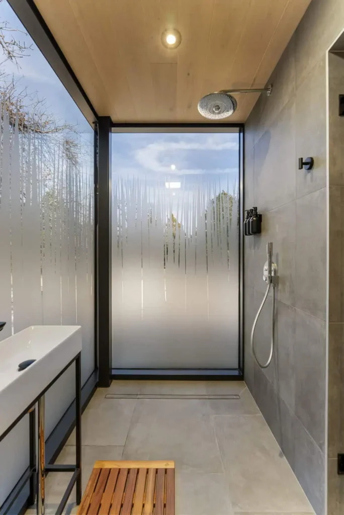 Modern shower area with frosted glass, rainfall showerhead, sleek sink, wooden bench, and natural light from large window, featured in Cosmic Cube accommodation at All Is Well.