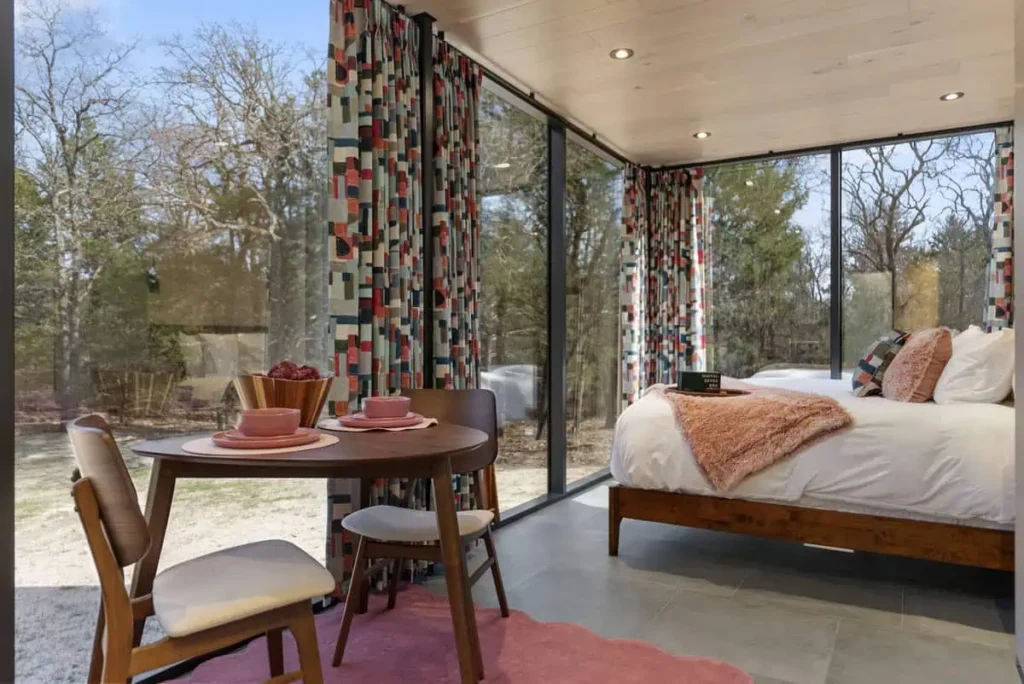 Cozy interior of the Cosmic Cube accommodation featuring a dining table with pink plates and a bowl of fruit, colorful curtains, a bed with a plush throw, and large windows showcasing the natural surroundings.
