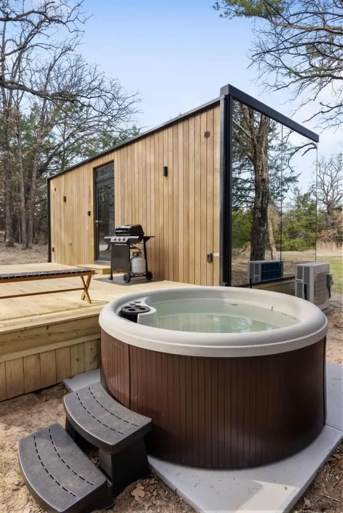 Hot tub on a wooden deck beside a modern mirror house, surrounded by trees, featuring a propane grill and outdoor seating.