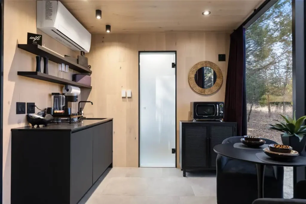 Modern interior of the Music Box accommodation featuring a kitchenette with a microwave, coffee machine, and sleek cabinetry, complemented by a dining area with decorative bowls and a view of nature outside.