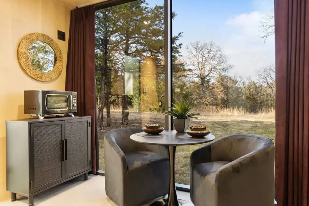 Cozy interior of the Music Box lodging at All Is Well Resort, featuring a round table with two chairs, a microwave, and a view of nature through large windows, complemented by a decorative mirror and potted plant.