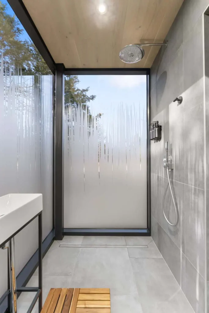 Modern outdoor shower with glass walls, rainfall showerhead, and wooden bench, showcasing a nature-inspired design for Music Box lodging at All Is Well Resort.