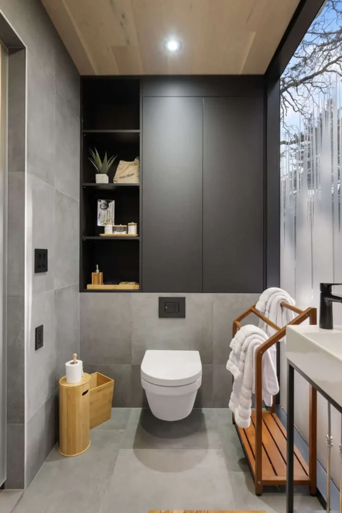 Modern bathroom interior featuring a wall-mounted toilet, wooden shelving with toiletries, and a towel rack, emphasizing a serene and stylish Zen Den retreat atmosphere.