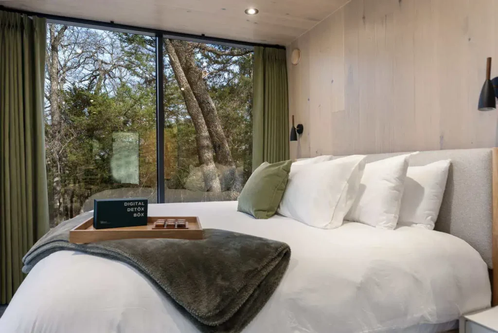 Cozy bedroom in the Mirror House featuring a king-sized bed with plush pillows, a digital detox box on a wooden tray, and large windows showcasing a serene natural view, emphasizing relaxation and wellness amenities.