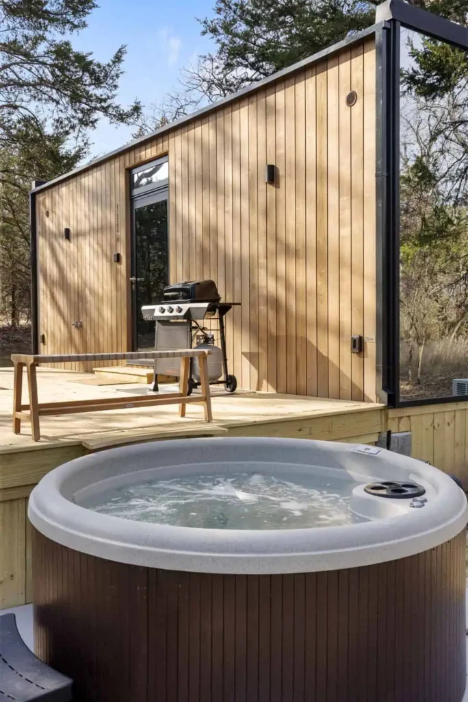 Hot tub in front of wooden cabin with grill and seating area, surrounded by trees, highlighting relaxation amenities at Mirror House Zen Den retreat.
