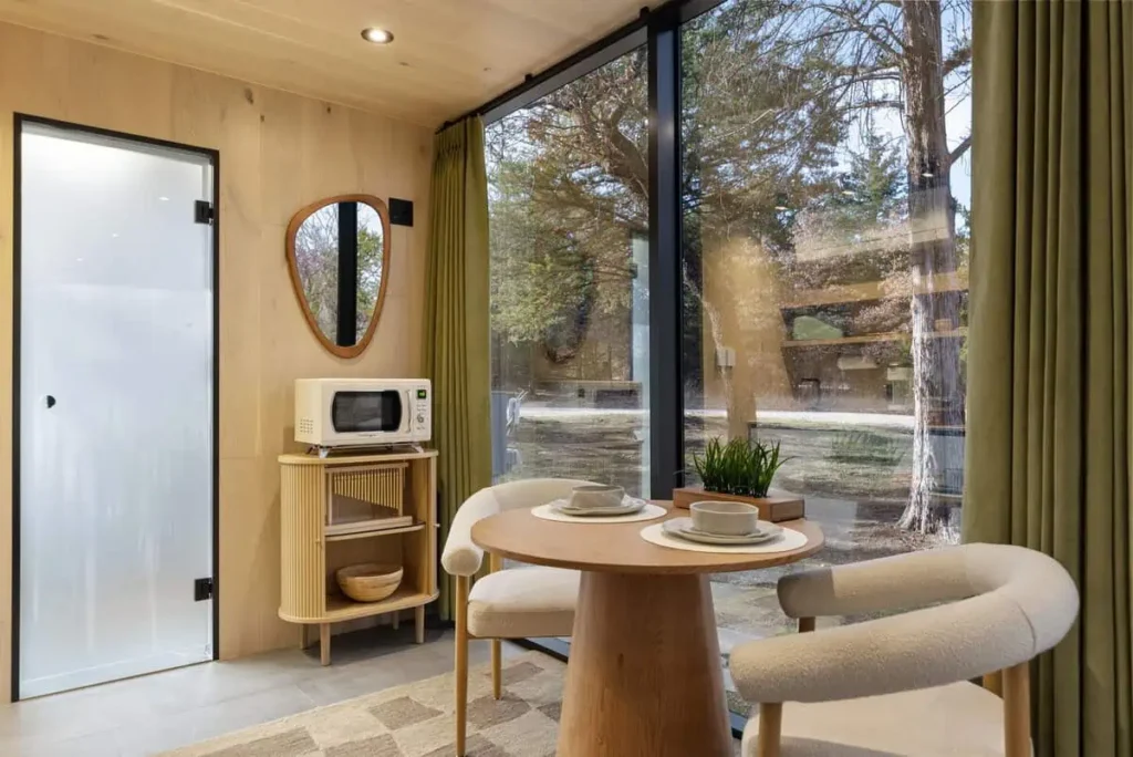 Interior of the Zen Den at All is Well St. Joe, featuring a modern table setting with two plates and cups, a microwave, and a large window revealing natural surroundings, emphasizing relaxation and comfort.