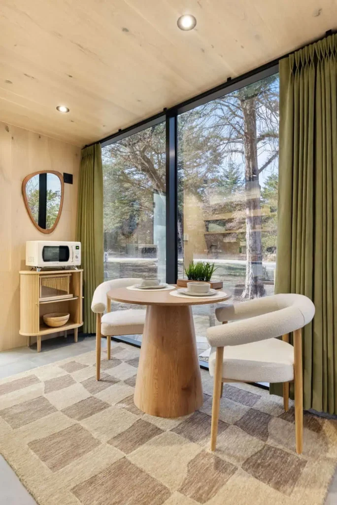 Interior of the Zen Den at All is Well St. Joe, featuring a round wooden table with two chairs, a microwave, and large windows with green curtains overlooking nature.