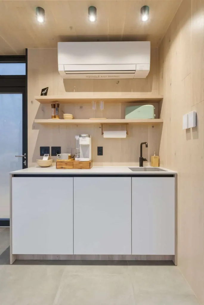 Modern kitchen area featuring air conditioning unit, minimalist cabinetry, wooden shelves with kitchenware, and coffee machine, highlighting amenities for the Zen Den retreat at All is Well St. Joe.