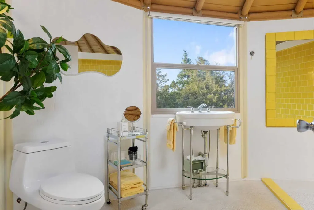 Bright and airy bathroom of the 'Hippy Chick' yurt, featuring a modern toilet, a vintage-style sink, yellow tiled accents, a potted plant, and a window with a view of nature.