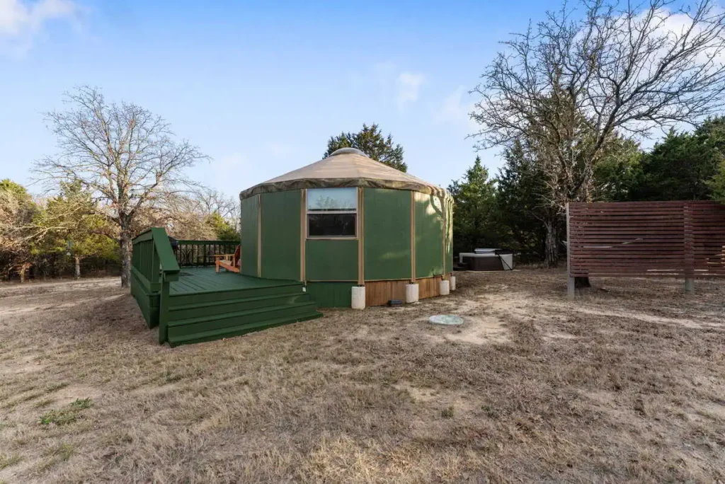Exterior view of the 'Hippy Chick' yurt at All Is Well Resort, featuring a green deck, surrounded by trees and a hot tub area, emphasizing unique glamping accommodations in nature.