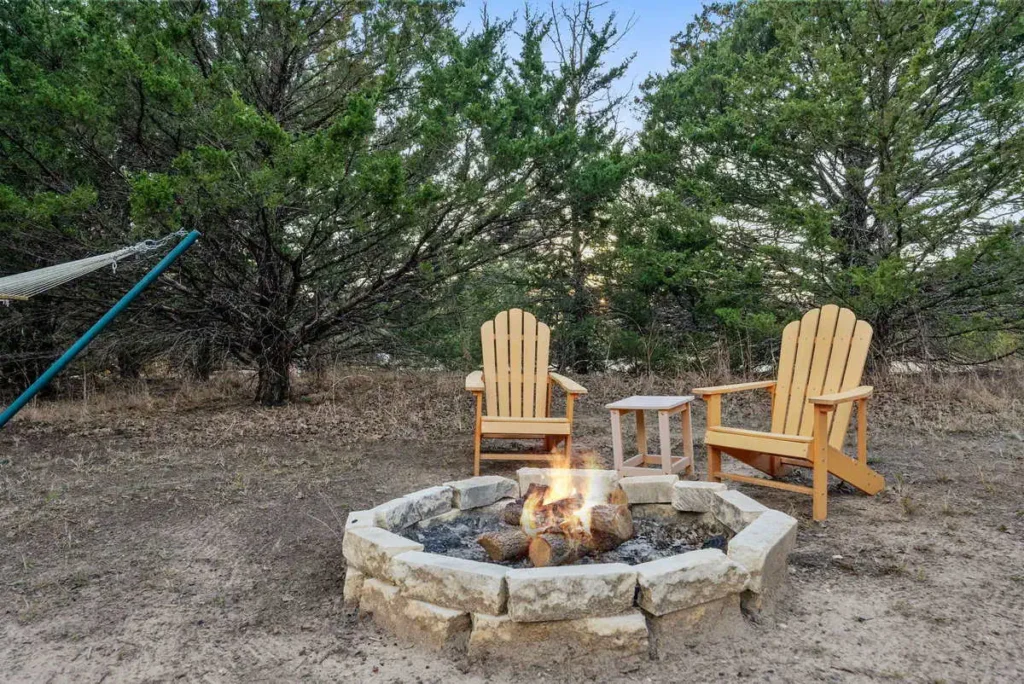Cozy fire pit area with two wooden Adirondack chairs, surrounded by trees and a hammock, creating a serene outdoor setting for relaxation at the Hippy Chick yurt accommodation.