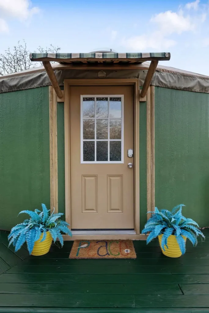 Hippy Chick yurt entrance featuring a green exterior, beige door, welcome mat, and vibrant blue potted plants on a wooden deck.