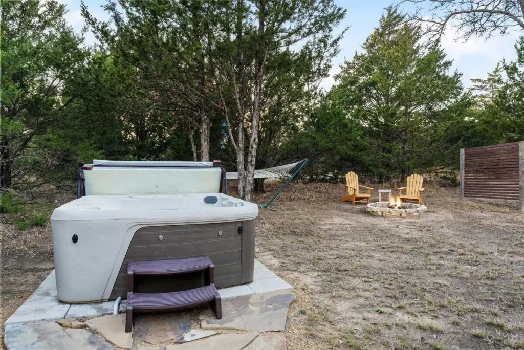 Hot tub in a natural setting near the 'Hippy Chick' yurt, surrounded by trees, hammock, and fire pit with seating.