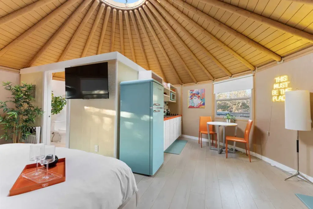 Interior of the 'Hippy Chick' yurt at All Is Well Resort, featuring a cozy bed with a tray, a turquoise refrigerator, a TV, dining area with orange chairs, and decorative plants, highlighting glamping amenities and whimsical design.