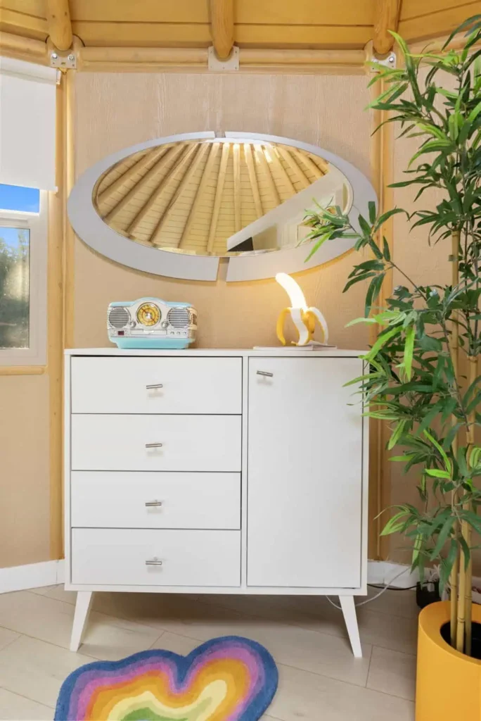 Interior of 'Hippy Chick' yurt featuring a white dresser, decorative mirror, colorful heart-shaped rug, and potted plant, emphasizing cozy and whimsical design elements for a unique glamping experience.